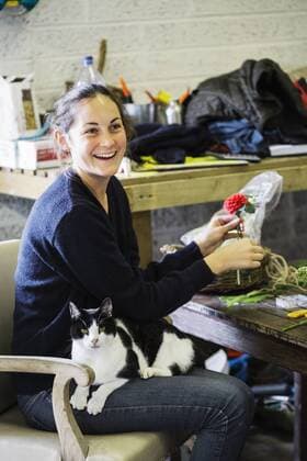 A woman in a flower barn, working on an arrangement with a cat sitting on her lap. A woman in a flower barn, working on an arrangement with a cat sitting on her lap.  22631
