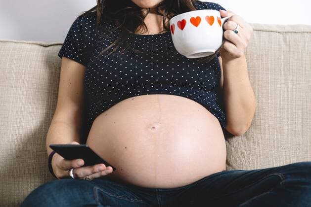 Pregnant woman showing her belly while sitting on the couch with cup of tea and smartphone model released Symbolfoto  GEMF01204