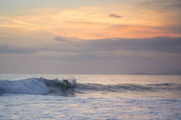 An orange colored sky accompanies a male surfer as he rides a small wave to the beach., Puerto Escondido Oaxaca Mexico Copyright: AndresxValencia VAAN000053