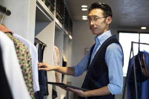 Japanese salesman with moustache wearing glasses standing in clothing store, looking at clothes on a rail, holding digital tablet. Japanese salesman with moustache wearing glasses standing in clothing store, looking at clothes on a rail, holding digital tablet.  43001