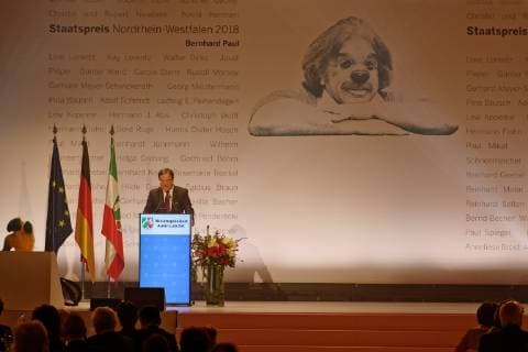 Award of the State Prize of the State of North Rhine-Westphalia 2018 to Bernhard Paul The picture shows the Minister President of the State of North Rhine-Westphalia on the speakers podium on the stage during his speech Guests can be seen in the foreground Behind Laschet a graphic by Bernhard Paul as clown Cologne North Rhine-Westphalia Germany is emblazoned on the back wall