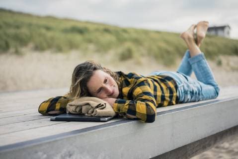 IMAGO Creative: Internationaler Frauenmonat, Gesundheit, Frauen in der Menopause Mature woman with laptop, lying on boardwalk at the beach, relaxing model released Symbolfoto  MOEF01646