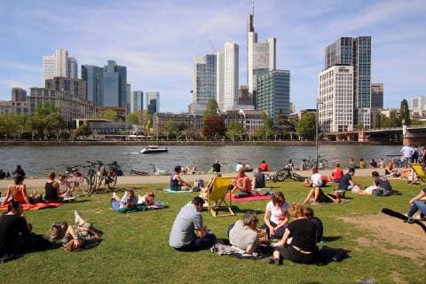 Spring day on Easter Monday in Frankfurt Sun lovers enjoy the warm temperatures and the sunbeams under a blue sky on a sunbathing lawn on the banks of the Main in front of the skyscrapers of the banking district s in downtown Hessen Germany
