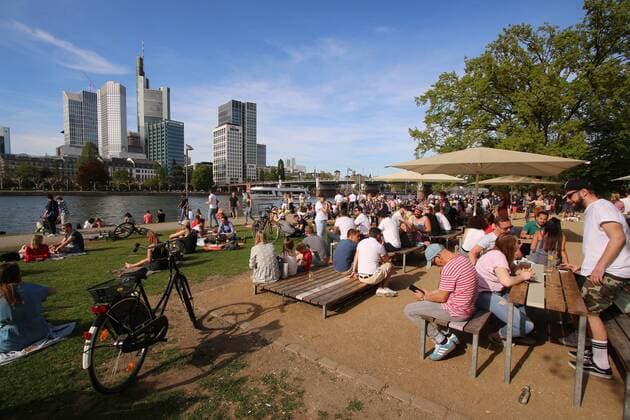 Spring day on Easter Monday in Frankfurt Sun lovers enjoy the warm temperatures and the sunbeams under a blue sky on a sunbathing lawn with beer garden on the banks of the Main in front of the skyscrapers of the banking district s in downtown Hessen Germany