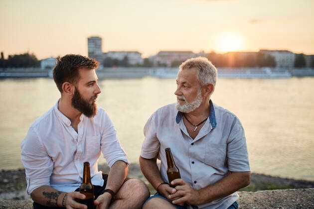 IMAGO Creative: Vaterschaft Father and adult son sitting on a wall at the riverside at sunset drinking a beer model released Symbolfoto  ZEDF02557