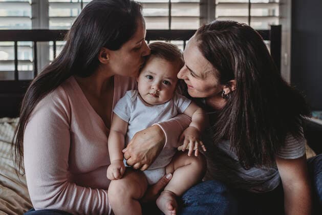 Two happy moms cuddling and kissing baby girl on bed near window La Mesa, CA CR_KESM191114A-213299-01