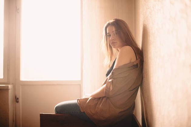 Portrait of young woman sitting on headboard of bed by window at home CR_DMBE200115-273232-01