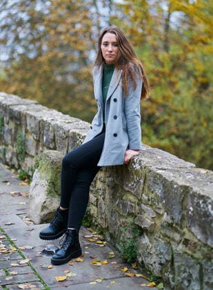 portrait of a young Caucasian woman with brown hair on a stone bridge with the green of the trees in the background. Dressed in a gray jacket, green sweater, dark pants and military boots Pamplona, Navarre, Spain ,model released, Symbolfoto CR_SEBO200124B-290044-01