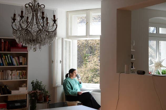 Junge Frau (model released) liest ein Buch an einem offenen Fenster in ihrer Wohnung. Studium zu Hause während der Corona-Krise. MR=YES