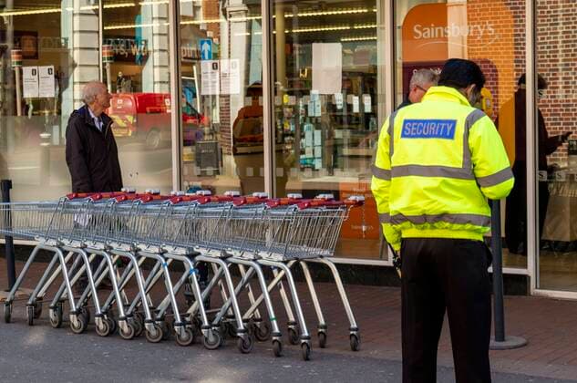 Security officer outside a Winchester supermarket. Allowing only one customer to enter at a time during Coronavirus epidemic, Winchester, Hampshire, England, UK. 65197_02_4421_027
