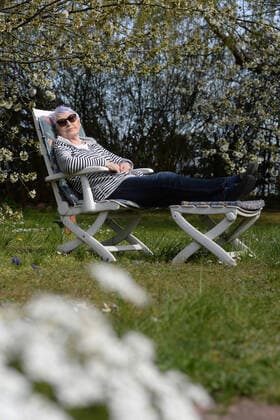 Ältere Frau (model released) entspannt sich auf einer Gartenliege in der Sonne in einem Garten mit blühenden Kirschbäumen MR=YES