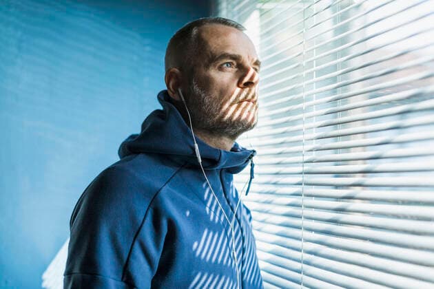 Pensive man with earbuds looking out of venetian blind window model released Symbolfoto property released MCVF00280