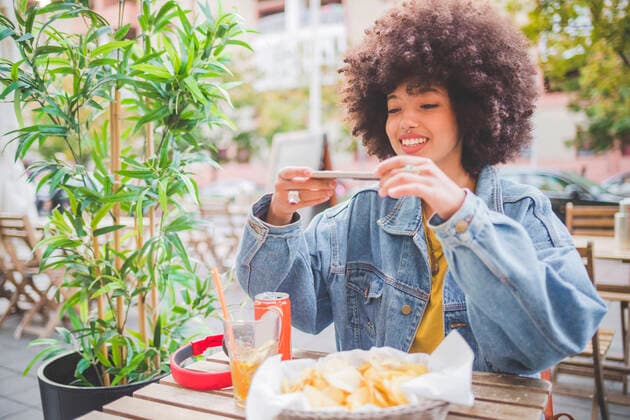 Smiling young woman with afro hairdo taking smartphone picture at an outdoor cafe in the city model released Symbolfoto MEUF00248