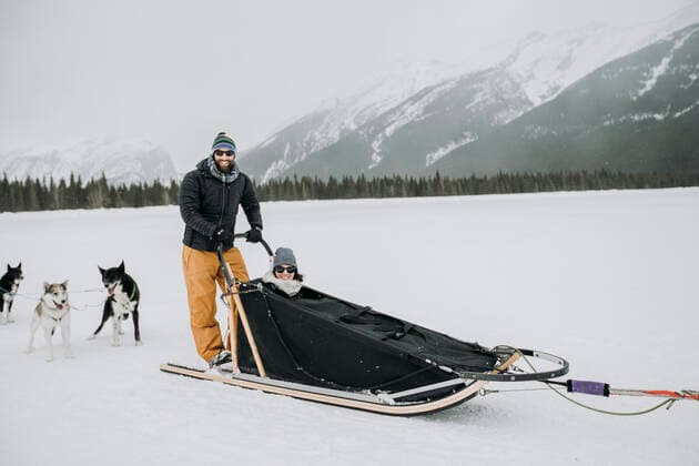 IMAGO Creative: Alternativer Wintersport man and woman couple sit and stand in dogsled in canadian rockies Banff, AB, Canada CR_SSNF200422A-341536-01,model released, Symbolfoto