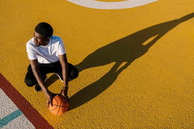 Thoughtful young man holding basketball crouching on sports court during sunny day model released Symbolfoto EGAF00294