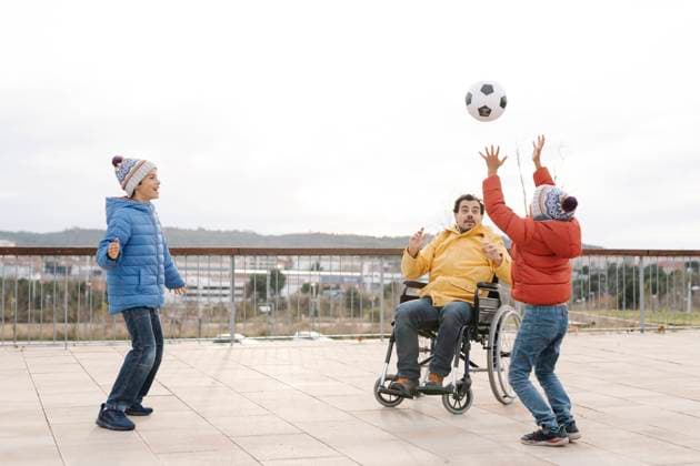 Man sitting on wheelchair throwing ball towards playful sons in park model released Symbolfoto JCZF00116