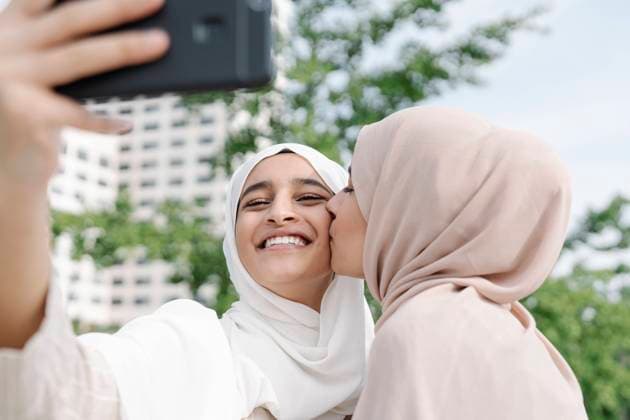Muslim woman kissing sister while taking selfie through smart phone in city model released Symbolfoto JCZF00145