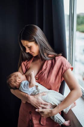 IMAGO Creative: Mutterschaft Mother breastfeeding baby boy while standing by window at home model released Symbolfoto OCMF01435