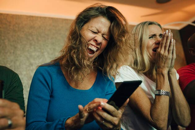 Happy woman cheering by friends while watching sports on smart phone in living room, MA82792, Sweden, Stockholm, model released, property released Copyright: xMaskotx