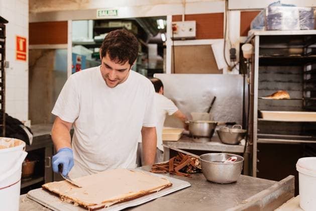 Smiling man in white uniform and latex gloves smearing cream on cake base while working in bakery kitchen, Model released, Property released Copyright: xAlbertoxMenendezx MR