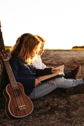 Side view of adorable little girl reading interesting story to younger brother while sitting together under tree with ukulele guitar in summer day in countryside Copyright: xAngelxSantamariax Model Released