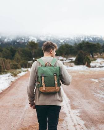 IMAGO Creative: Jahreszeit Winter, Spaziergang Back view of young male traveler in sweater with backpack walking on dirt rural road in cloudy winter day with green forest and snow on roadsides, Model released Copyright: xCarlosxForteax MR