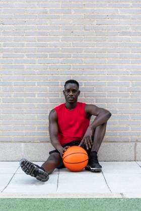 Confident African American male basketball player in sportswear sitting with ball on stone border on playground and looking at camera Copyright: xDavidxEspejox Model Released