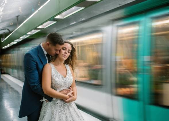 Young bride in white wedding dress standing with closed eyes while groom standing behind and hugging with moving train on blurred background at subway Copyright: xGabrielxTrujillox Model Released