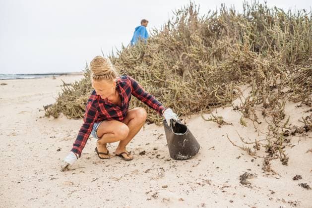 Positive hipster woman in casual clothes and gloves collecting trash into bag while squatting on deserted beach Copyright: xGabrielxTrujillox Model Released