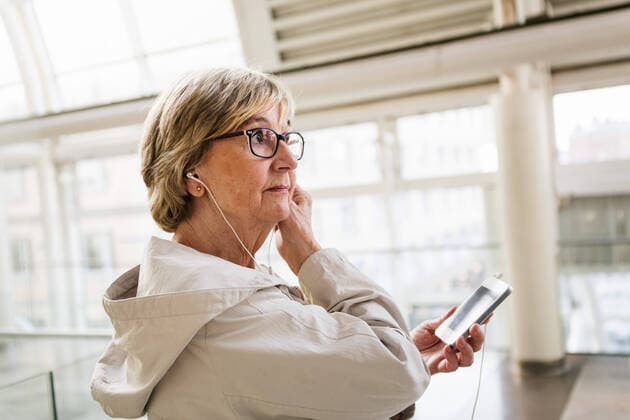 IMAGO Creative: Stadt-Dichotomien Side view of senior woman adjusting in-ear headphones while standing with smart phone at railroad station , model released Copyright: xKentarooxTrymanx , MA62940
