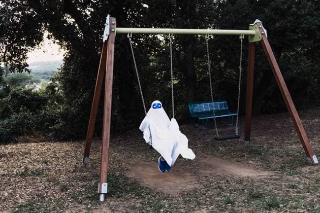 Unrecognizable kid in white ghost costume swinging and playing on playground during Halloween holiday, Model released Copyright: xIvanxPujolx MR