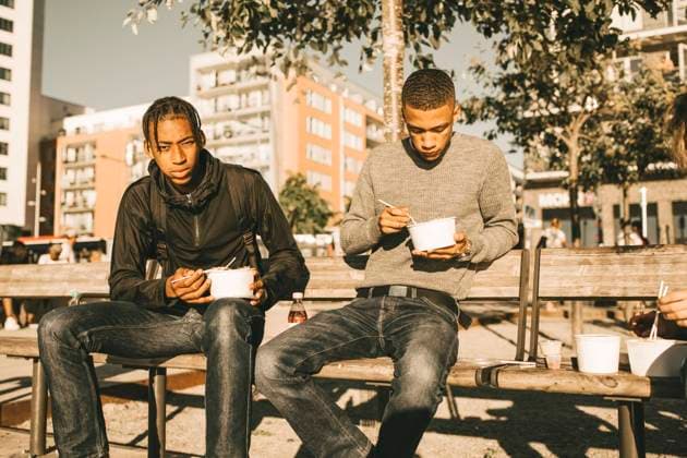 Portrait of serious teenage boy holding take out food while sitting by friend on bench in city during sunny day , model released Copyright: xMaskotx , MA69393