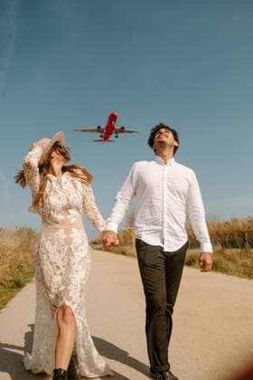 Happy young man in stylish outfit holding hand of woman in white wedding dress while strolling together on country road against blue sky with flying airplane, Model released Copyright: xJakexJakabx MR