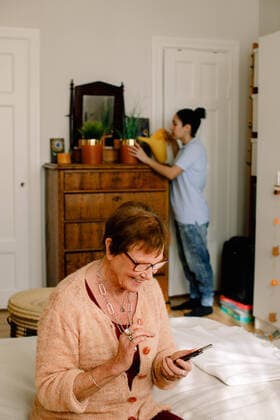 IMAGO Creative: Zivildienst und ehrenamtliches Engagement Smiling retired senior woman using smart phone while volunteer watering plants in bedroom at nursing home , model released, property released Copyright: xMaskotx , MA70819