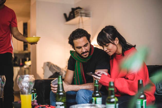 IMAGO Creative: Sport Start-Up Male and female friends using smart phone while watching match at home , model released, property released Copyright: xMaskotx , MA77385