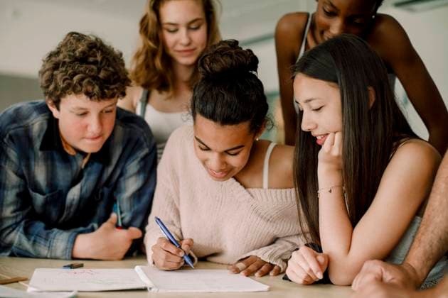 IMAGO Creative: Bildung, Schreiben Smiling teenage girl writing in book while sitting amidst friends in classroom , model released, property released Copyright: xMaskotx , MA77522