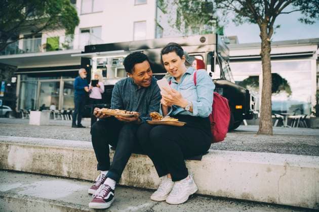 Smiling friends sharing smart phone while sitting with meal against food truck in city Sweden, Stockholm, model released, property released Copyright: xMaskotx , MA81566