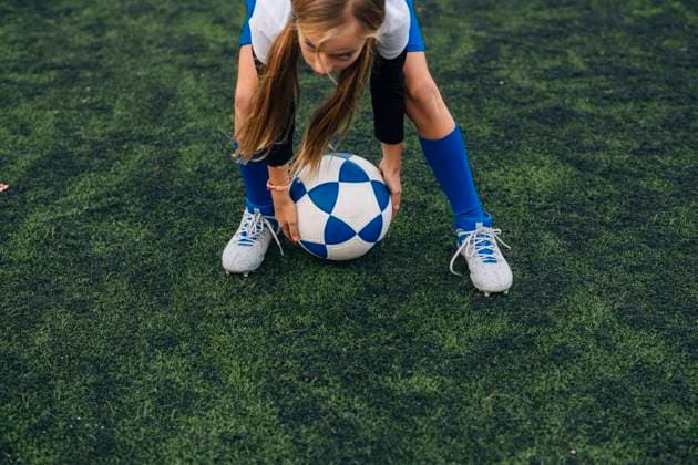 teenage girl in white and blue uniform and cleats running and preparing to kick ball while playing soccer alone on green field in contemporary sports club, Model released Copyright: xJosexLuisxCarrascosax MR