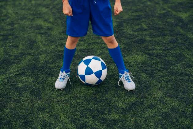 IMAGO Creative: NGO, Ausbildung f?r Kinder High angle of faceless child in blue shorts and cleats with ball standing alone on green court while playing football at modern stadium, Model released Copyright: xJosexLuisxCarrascosax MR