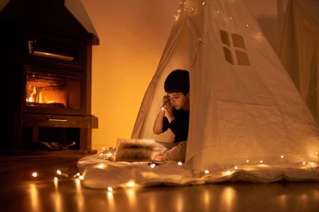 IMAGO Creative: Herbst, Gemütliches Zuhause Focused little kid in casual wear reading book while lying in kids play tent with flashlight and garland around near fireplace in apartment, Model released Copyright: xJuanxGarciaxRisquezx MR