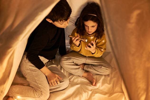 IMAGO Creative: Herbst, Gemütliches Zuhause Curios boy watching through spyglass while sitting with crossed legs near dreamy little sister in casual wear taking garland in evening in flat, Model released Copyright: xJuanxGarciaxRisquezx MR