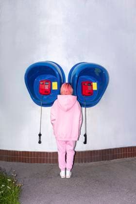 IMAGO Creative, Lifestyle, Barbiecore Rear view of young woman with pink hair wearing pink hooded shirt standing in front of telephone booths model released Symbolfoto VPIF03277