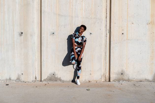 IMAGO Creative: Neue Aussichten Cheerful confident African American female athlete in flowered sport clothes and white sneakers looking away with interest and laughing while standing alone leaning on concrete wall in sunbeams and resting after training in city, Model released Copyright: xManuelxOrtsx MR