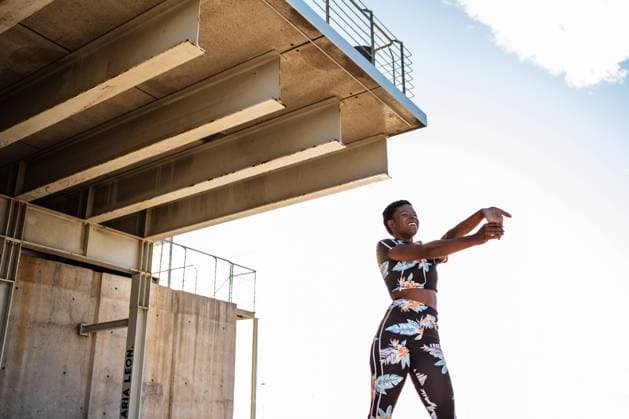 IMAGO Creative: Clickbait, Sport und Fitness From below focused African American adult woman in flowered sportswear stretching hand muscles while standing alone and warming up before training among urban environment in sunny day, Model released Copyright: xManuelxOrtsx MR