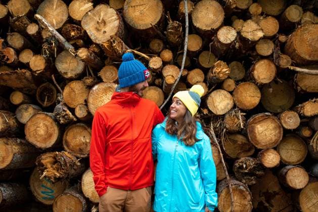 IMAGO Creative: Landleben - Alfresco, Drau?en Happy young man and woman in warm outerwear and hats embracing and looking at each other while standing near heap of cut tree logs in forest Copyright: xMarinaxTobaruelax Model Released
