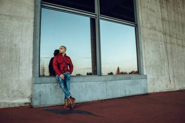 Confident young unshaven male in red warm jacket and jeans looking away while standing and leaning against a big window Copyright: xMartaxMartinezx Model Released
