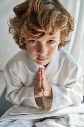 Thoughtful young boy in white kimono sitting with legs crossed and hands together while practicing meditation looking at camera during training judo at home Copyright: xPhilippexDegrootex Model Released