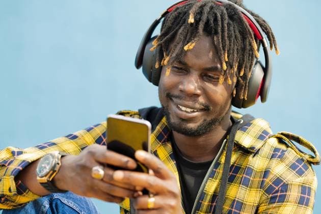 IMAGO Creative: Bildung, Schreiben portrait of a young black man with a headphones writing on his smart phone with a blue wall in the background, technology and lifestyle concept, copy space for text, Model released Copyright: xRaulxMelladox MR
