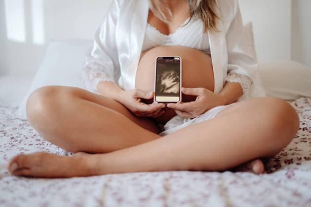 Crop unrecognizable pregnant female demonstrating picture of baby hans from ultrasound scan on smartphone while sitting in bra and open shirt on bed with crossed legs, Model released Copyright: xSantixMiquelx MR