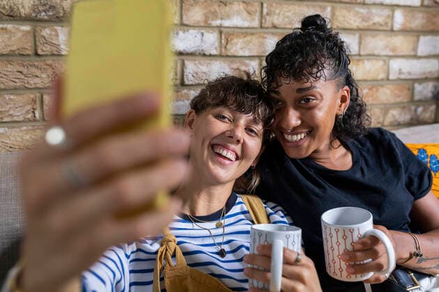 Young lesbian couple with tea cup taking selfie through mobile phone while sitting at home model released Symbolfoto  WPEF03599